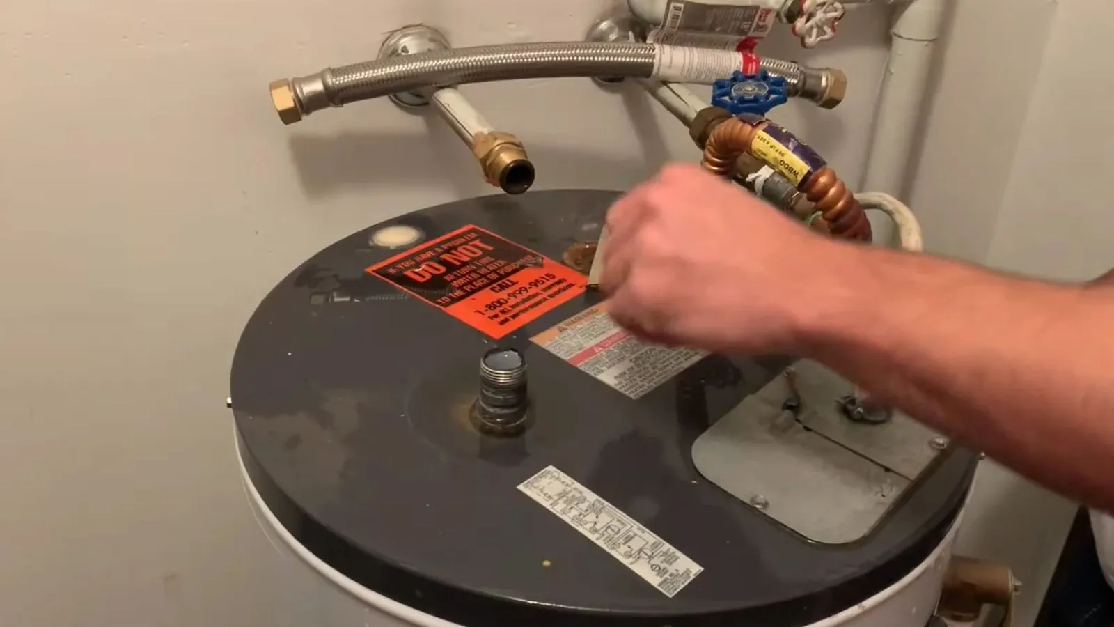 Hand at the top of a water heater tank beside the supply connections and warning label